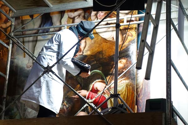 A skilled craftsman wearing a white lab coat is restoring a large, old painting.