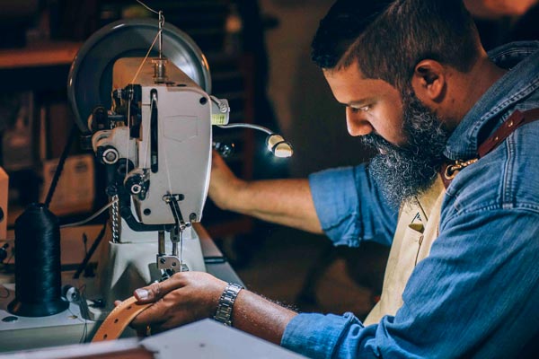 A professional man with a beard and wearing a blue denim shirt is working at a sewing machine making leather belts.